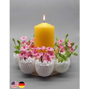 May include: A crocheted Easter egg candle holder with pink flowers and green leaves surrounding a yellow candle.