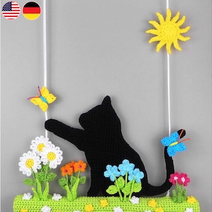 Può includere: Un gatto nero all'uncinetto con una lunga coda appesa a uno spago bianco. Il gatto è circondato da fiori all'uncinetto in tonalità di bianco, rosa, blu e giallo. Ci sono anche due farfalle all'uncinetto, una gialla e una blu.