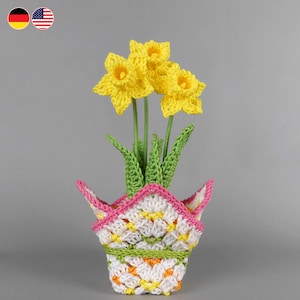 May include: A crocheted daffodil arrangement in a decorative pot. The yellow flowers have green stems and leaves. The pot is white with a colorful pattern, a pink border, and a green band.
