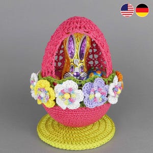 May include: A crocheted Easter egg basket with a pink base and a green rim. The basket is filled with a chocolate bunny and colourful Easter eggs. The basket is decorated with crocheted flowers in yellow, white, and purple.