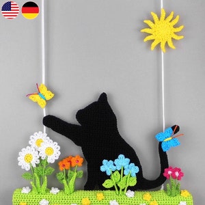 May include: A black crocheted cat with a long tail hanging from a white string. The cat is surrounded by crocheted flowers in shades of white, pink, blue, and yellow. There are also two crocheted butterflies, one yellow and one blue.