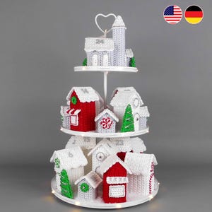 May include: A three-tiered white metal stand displaying a collection of crocheted miniature houses. The houses are red, white, and grey, featuring snowy roofs, small green trees, and numbered doors. The stand is illuminated by small white lights.
