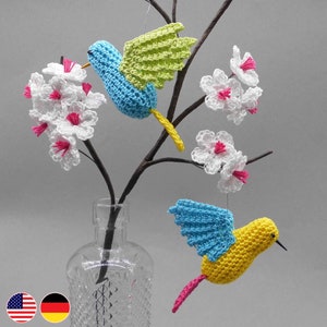 May include: Crocheted hummingbird ornaments in blue, yellow, and pink, with green wings, hang from a branch with white and pink flower accents. The arrangement is displayed in a clear glass vase, creating a whimsical, nature-inspired decor.
