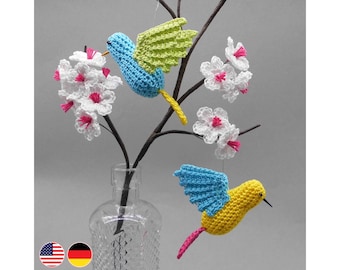 May include: Crocheted hummingbird ornaments in blue, yellow, and pink, with green wings, hang from a branch with white and pink flower accents. The arrangement is displayed in a clear glass vase, creating a whimsical, nature-inspired decor.
