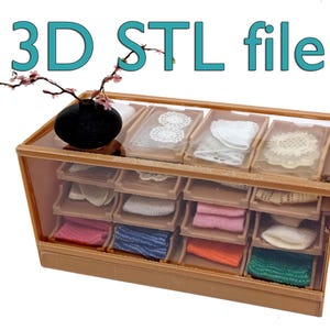 May include: A miniature display case with multiple compartments, filled with various lace doilies and colorful spools of thread. The text "3D STL file" is displayed in teal. A small black vase with a sprig of pink flowers sits on top.