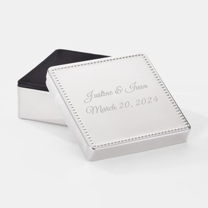 May include: A square silver-toned metal keepsake box with a beaded border on the lid. The lid is engraved with the text Justine & Ivan and the date March 20, 2024. The box is open, revealing a black interior lining within the silver-toned base.