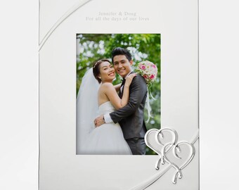 May include: Silver picture frame with a white mat and a photo of a bride and groom. The frame has a silver ribbon design and two intertwined hearts at the bottom. The inscription reads "Jennifer & Doug For all the days of our lives."