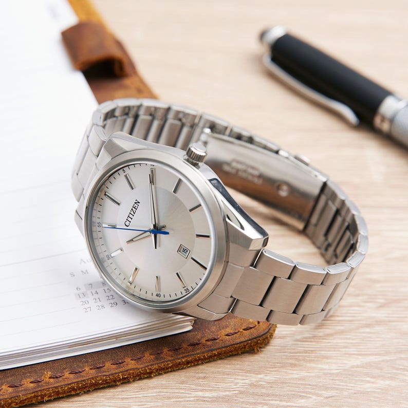 Puede incluir: Un reloj Citizen plateado con una correa plateada descansa sobre un bloc de notas. El reloj tiene una esfera plateada con agujas azules y la palabra "CITIZEN". Un bol&iacute;grafo negro est&aacute; en el fondo. Un accesorio elegante.