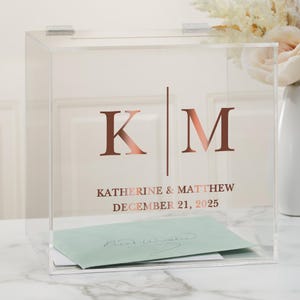 May include: Clear acrylic wedding card box with a slot on the top. The box has a rose gold inscription that reads "K | M Katherine & Matthew December 21, 2025".