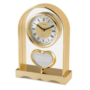 Puede incluir: Un reloj de mesa dorado con un marco de vidrio transparente y una placa en forma de coraz&oacute;n. La esfera del reloj tiene n&uacute;meros romanos y las palabras "Danbury Quartz". La placa dice "Celebrating 50 Years Ed & Molly Warner Established in 1973".