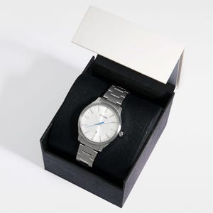 Puede incluir: Un reloj Citizen plateado con una correa de metal a juego, presentado en una caja negra con tapa blanca. El reloj tiene una esfera clara con manecillas plateadas y detalles azules. Dise&ntilde;o cl&aacute;sico.