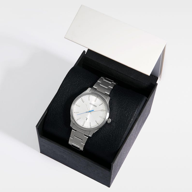 Puede incluir: Un reloj Citizen plateado con una correa de metal a juego, presentado en una caja negra con tapa blanca. El reloj tiene una esfera clara con manecillas plateadas y detalles azules. Dise&ntilde;o cl&aacute;sico.
