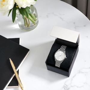 Puede incluir: Un reloj de pulsera plateado con esfera clara y correa met&aacute;lica se muestra en una caja negra con tapa blanca. Un bol&iacute;grafo dorado y dos cuadernos negros est&aacute;n a la izquierda. Un jarr&oacute;n con flores blancas est&aacute; en el fondo.
