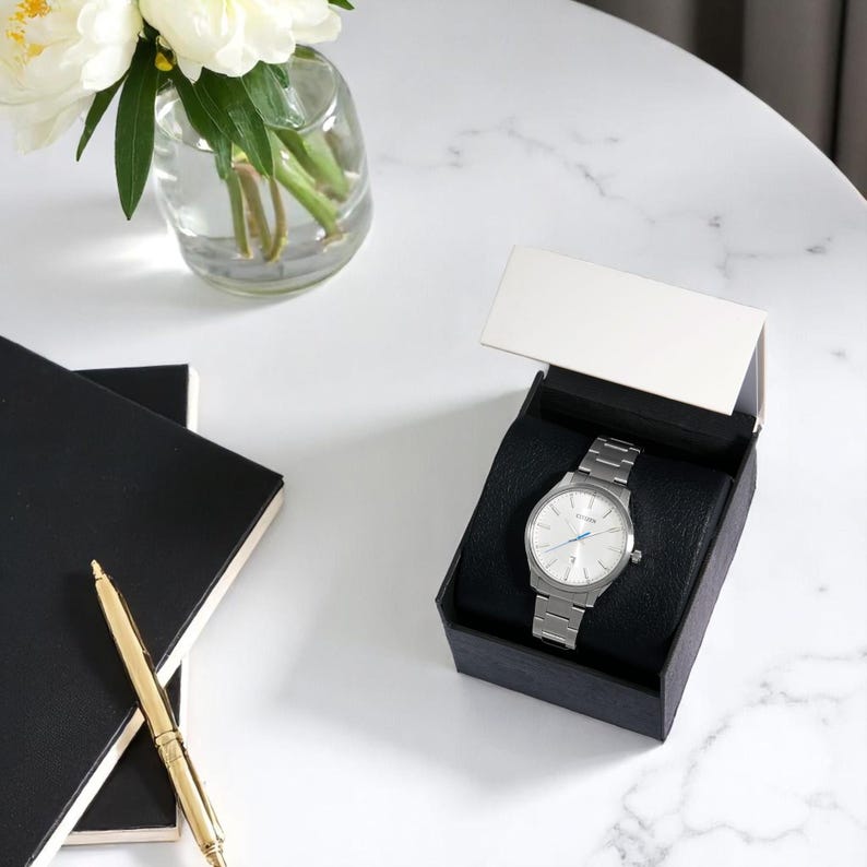 Puede incluir: Un reloj de pulsera plateado con esfera clara y correa met&aacute;lica se muestra en una caja negra con tapa blanca. Un bol&iacute;grafo dorado y dos cuadernos negros est&aacute;n a la izquierda. Un jarr&oacute;n con flores blancas est&aacute; en el fondo.