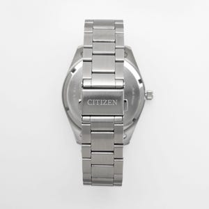 Puede incluir: Vista trasera de un reloj Citizen de acero inoxidable. La correa del reloj es una serie de eslabones rectangulares. El cierre est&aacute; grabado con el nombre de la marca "CITIZEN". La esfera del reloj es visible, con texto y una peque&ntilde;a corona.