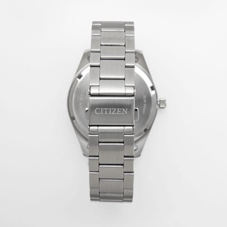 Puede incluir: Vista trasera de un reloj Citizen de acero inoxidable. La correa del reloj es una serie de eslabones rectangulares. El cierre est&aacute; grabado con el nombre de la marca "CITIZEN". La esfera del reloj es visible, con texto y una peque&ntilde;a corona.