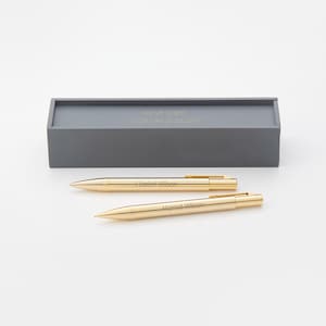 May include: Two gold-colored pens with the name "Harriet Wilson" engraved on them. The pens are in a gray box with the text "Harriet Wilson 2023 MBA Graduate" on the top.