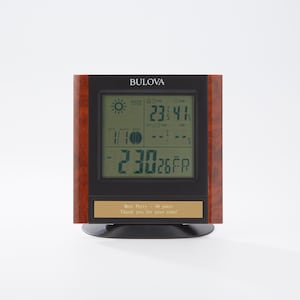 May include: A Bulova digital weather station with a black display and a wood-grain frame. The display shows the time, date, and weather information. A gold plaque at the bottom reads "Bret Perry - 40 years. Thank you for your time!"