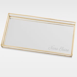May include: A rectangular glass tray with a gold metal frame. The tray has a mirrored surface and is engraved with the text "Nana Elaine".