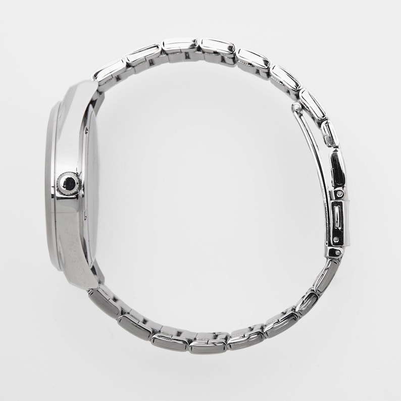 Puede incluir: Vista lateral de un reloj de pulsera plateado con una correa de metal con eslabones. La esfera del reloj es redonda y la correa est&aacute; compuesta por peque&ntilde;os eslabones rectangulares. El reloj est&aacute; sobre un fondo blanco.