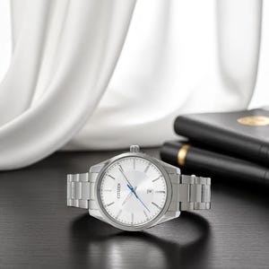 Puede incluir: Un reloj Citizen plateado con esfera blanca y agujas azules. El reloj tiene una correa de acero inoxidable y una pantalla de fecha. El reloj est&aacute; sobre una superficie negra con una tela blanca en el fondo.