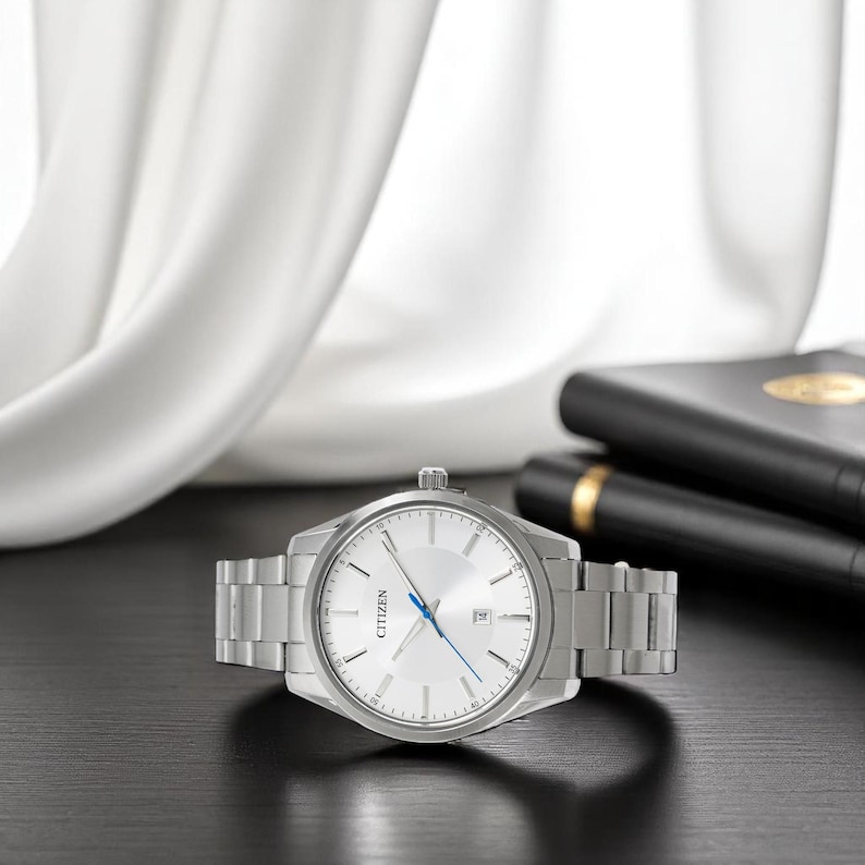 Puede incluir: Un reloj Citizen plateado con esfera blanca y agujas azules. El reloj tiene una correa de acero inoxidable y una pantalla de fecha. El reloj est&aacute; sobre una superficie negra con una tela blanca en el fondo.