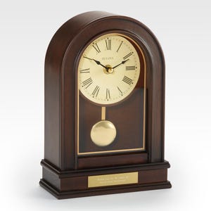 May include: A dark brown wooden Bulova mantel clock with a gold-colored clock face and pendulum. The clock has Roman numerals and a small plaque at the base with text. The clock is a classic, decorative item.