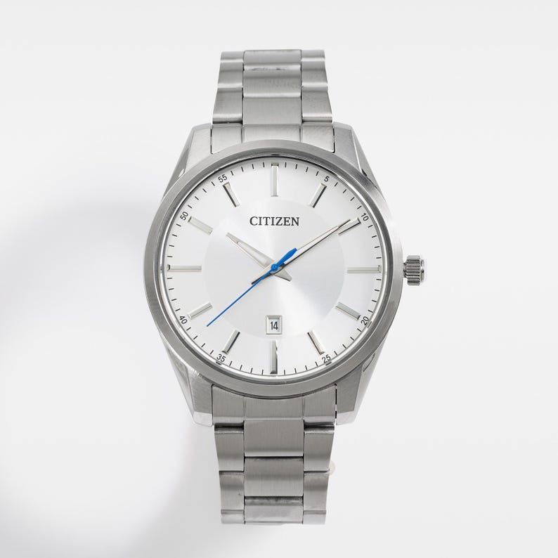 Puede incluir: Un reloj Citizen plateado con esfera blanca, agujas azules y visualizaci&oacute;n de fecha a las 3. El reloj tiene una correa de acero inoxidable y un bisel redondo. El reloj tiene un dise&ntilde;o cl&aacute;sico.