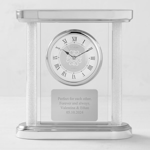 May include: A silver tabletop clock with a clear glass base and a silver face. The clock has a white background with black Roman numeral hour markers and black hands. The clock face also has the words "DANBURY" and "QUARTZ" printed on it. The clock has a silver plaque on the bottom with the text "Perfect for each other. Forever and always. Valentina & Ethan 05.10.2024".