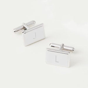 May include: A pair of silver cufflinks with rectangular faces. The cufflinks are engraved with the letters "J" and "L".