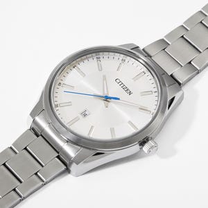 Puede incluir: Un reloj Citizen plateado con esfera blanca, visualizaci&oacute;n de fecha y segundero azul. El reloj tiene una correa de acero inoxidable y un bisel plateado. El reloj est&aacute; en &aacute;ngulo, mostrando la esfera y parte de la correa.