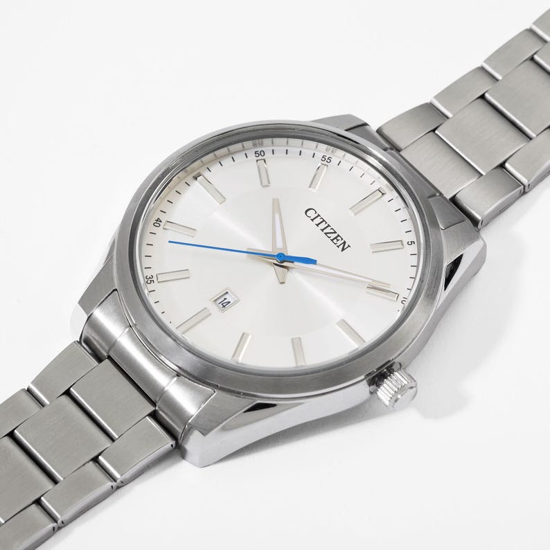 Puede incluir: Un reloj Citizen plateado con esfera blanca, visualizaci&oacute;n de fecha y segundero azul. El reloj tiene una correa de acero inoxidable y un bisel plateado. El reloj est&aacute; en &aacute;ngulo, mostrando la esfera y parte de la correa.