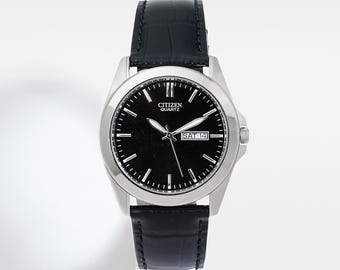 May include: A Citizen Quartz wristwatch with a black dial and silver-tone case. The watch features a black leather band and displays the day and date. The watch face has white hour markers and hands.