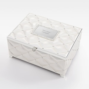 May include: A white, rectangular jewelry box with a quilted pattern and silver trim. The box features a rectangular plaque with the inscription "Parker Eleanor Cunningham" and is adorned with rhinestones. A decorative clasp secures the lid.