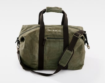 May include: Olive green duffel bag with brown accents, featuring a top handle, shoulder strap, and a front pocket. The name "Jackson" is embroidered in white on the front of the bag. The bag has a zipper closure.