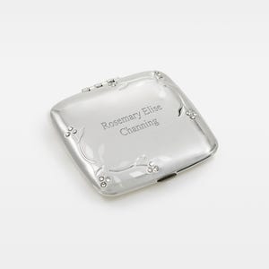 May include: A silver compact mirror with rounded corners and a hinged lid. The surface is engraved with the name "Rosemary Elise Channing" and features a delicate floral design along the edges. The compact is closed and reflects the surrounding light.
