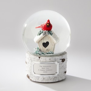 May include: A white snow globe with a red cardinal perched on a birdhouse. The base is a birch tree log with a silver plaque that reads "The Mavericks Wish You A Happy Holiday! 2023".