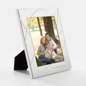 May include: A silver picture frame with a white mat and a decorative design. The frame has a stand and is displaying a photo of a family of three in a park. The text "The Wellesley Family" is printed on the bottom of the frame.