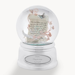 May include: A clear glass snow globe featuring a scroll with the text The Lord bless you and keep you, a silver butterfly, and pink floral vines. The silver base includes an oval plaque engraved with the text Christina, May faith always be your guide.