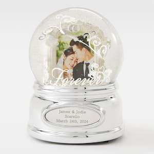 May include: A silver-colored snow globe with a photo of a couple inside. The globe has a decorative arch with the word "Forever" and the names "James & Jodie Scarello" and the date "March 24th, 2024" on the base.