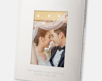 May include: A silver picture frame with a white mat and a photo of a couple in a wedding setting. The photo shows the couple looking at each other with a veil over the woman's head. The frame has an inscription that reads "Life together is just beginning October 10th, 2024".