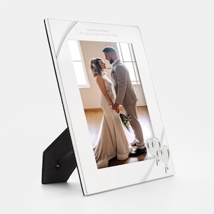 May include: A silver picture frame with a white ribbon and two silver hearts. The frame is standing on a black stand. The frame has a photo of a bride and groom inside. The text on the frame reads "Jennifer & Doug For all the days of our lives."