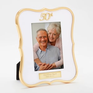 May include: White picture frame with a gold border and a gold 50th anniversary inscription. The frame has a photo of a couple inside and a gold plaque with the text "Tom & Carolyn Fitzgerald, October 12th, 1972".