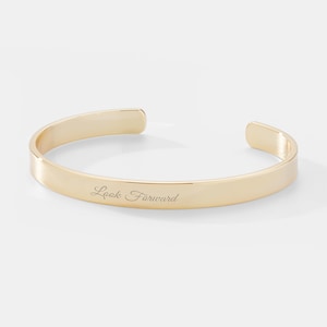 May include: Gold-toned bangle bracelet with the words "Look Forward" engraved on the front.