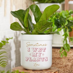 May include: White ceramic planter with the text "Sometimes I Wet My Plants" in pink and gold lettering. The name "Lori" is written in gold lettering below the text. A green plant is growing in the planter.