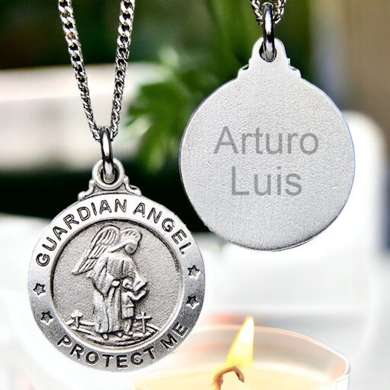 Puede incluir: Collar con colgante de &aacute;ngel de la guarda de plata con las palabras "Guardian Angel Protect Me" alrededor del borde. El colgante presenta una representaci&oacute;n de un &aacute;ngel con alas, sosteniendo un libro y de pie sobre un ni&ntilde;o. El collar tambi&eacute;n incluye una etiqueta de plata con el nombre "Arturo Luis" grabado en ella.