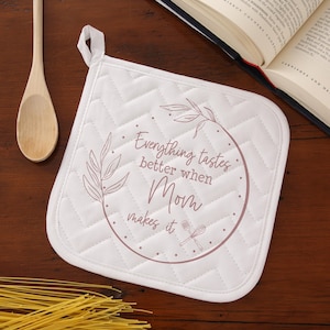 May include: White quilted pot holder with the text "Everything tastes better when Mom makes it" in a circular design. The design includes floral elements and a spoon and fork. A wooden spoon and a cookbook are in the background.