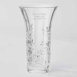 May include: A clear glass vase featuring an etched floral vine pattern around the exterior. The upper portion of the vase is engraved with the text: Forever and Always, Emly & Nick, May 27th, 2027. The vase has a flared rim and a thick base.