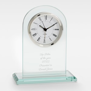 May include: A clear glass clock with a silver face and Roman numeral hour markers. The clock has an inscription that reads "Top Sales of the year 2023 Presented to Daniel Jones".