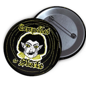 Puede incluir: Un botón negro con una ilustración amarilla y blanca de un vampiro y el texto "Compelled to Skate". El botón tiene un reverso de alfiler plateado y un diseño en espiral negro y amarillo.